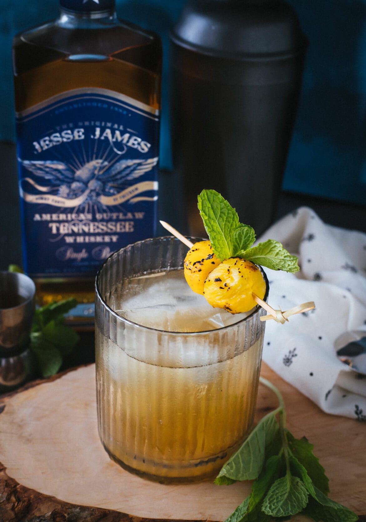 A glass of hula fashioned cocktail with pineapple syrup with mint and grilled pineapple balls for garnish, served on a wooden serving tray with a bottle of whiskey blurry in the background