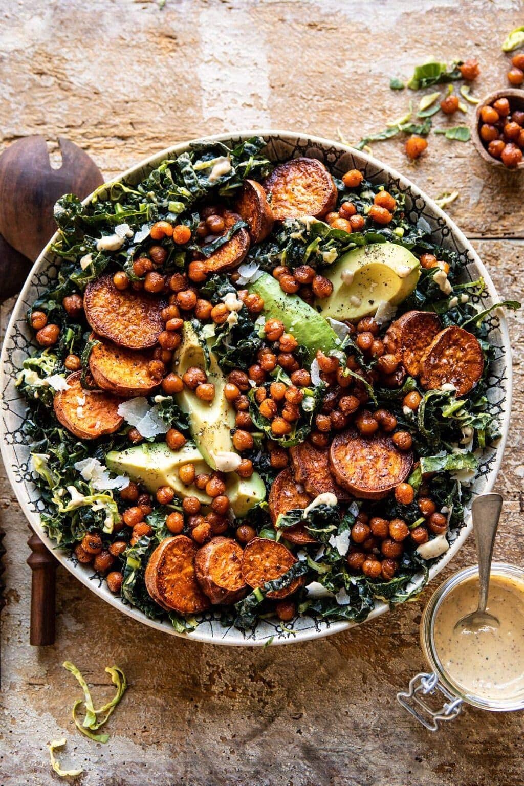Kale Caesar Salad with sweet potatoes, crispy chickpeas, shredded brussels sprouts, and avocado served on a plate with a glass of lemony parmesan tahini dressing on the side