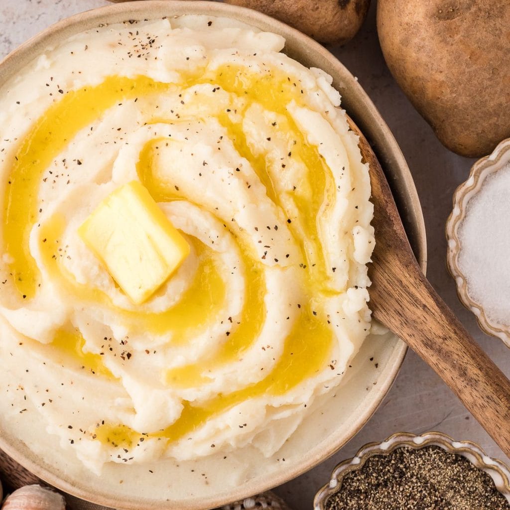 Brown bowl with mashed potatoes, butter slice, pepper, and wooden spoon.