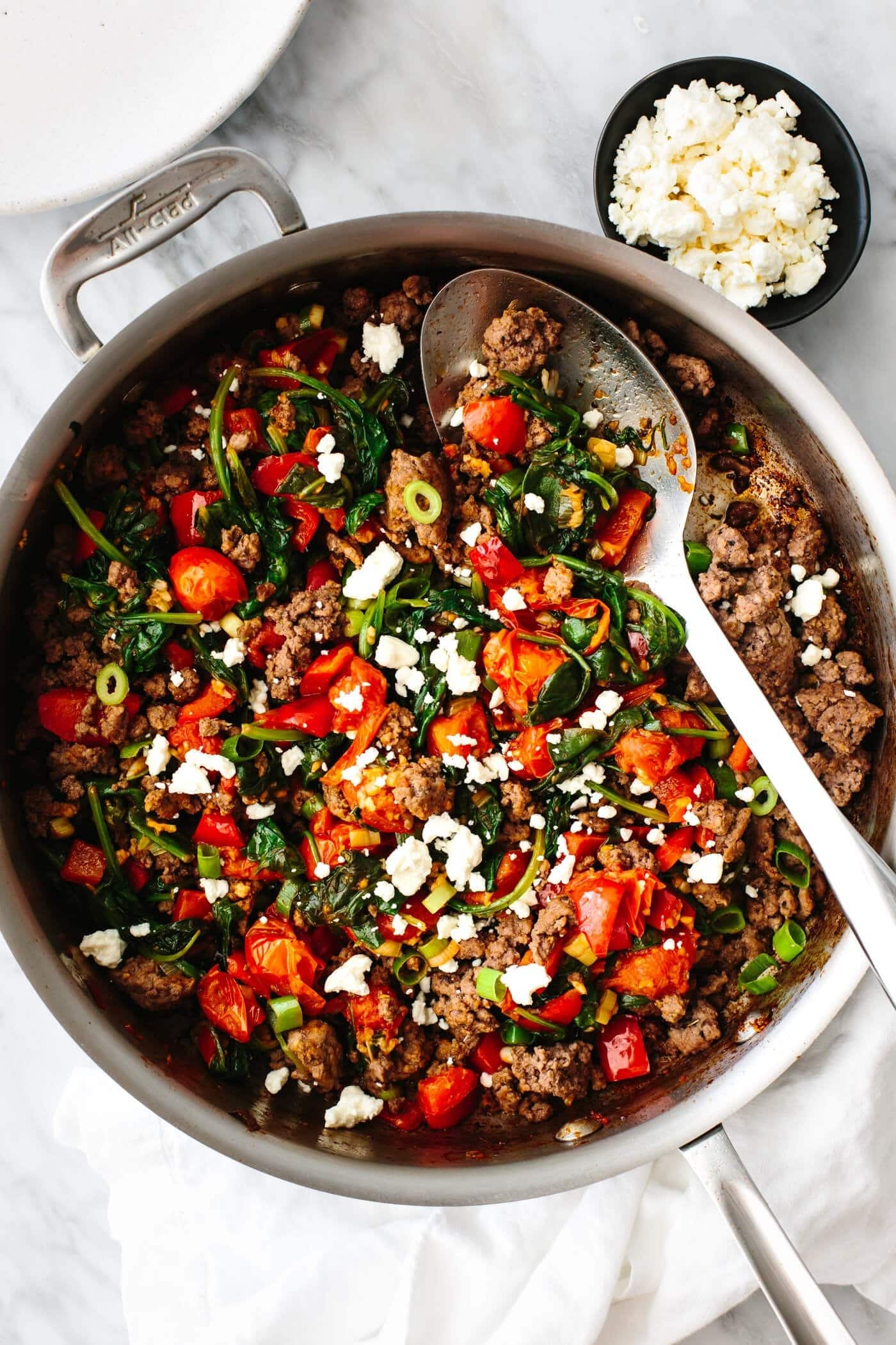 Mediterranean ground beef stir fry with spinach, tomatoes, ground beef, and scallions, and feta cheese in a bowl with a spoon.