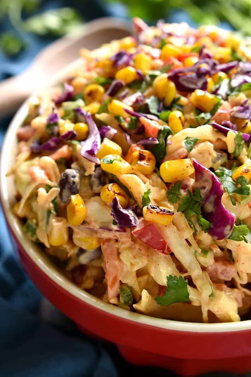 Coleslaw with corn, black beans, cilantro, red pepper and sliced jalapeno mixed with creamy dressing served on a red bowl. 