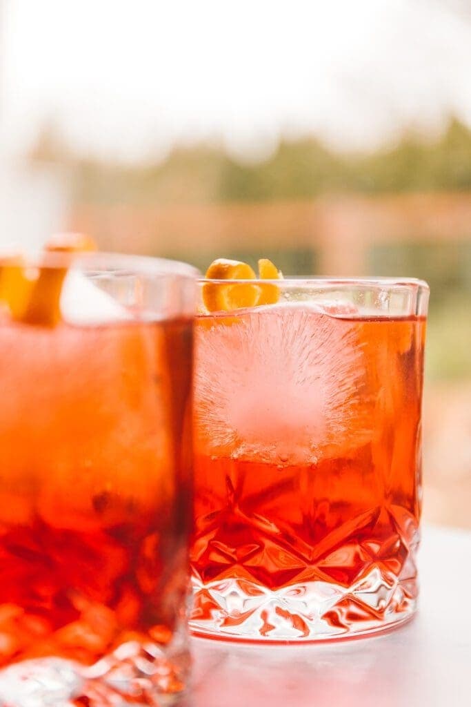 Two glasses of red orangey champagne cocktail with ice garnished with lemon peel.