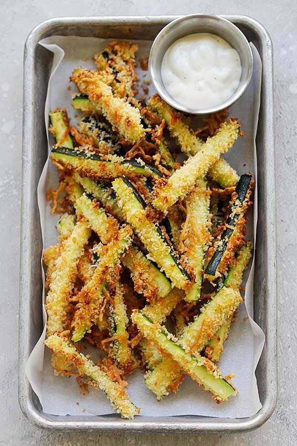 Breaded zucchini fries on a baking pan with liner served with white sauce. 