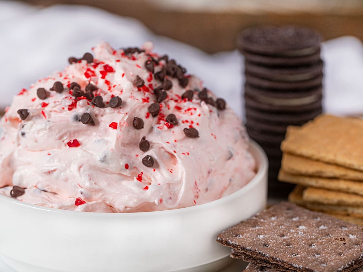 Christmas dessert dip on a bowl with chocolate chips, peppermint candy pieces, Cool Whip and cream cheese and different cracker beside. 