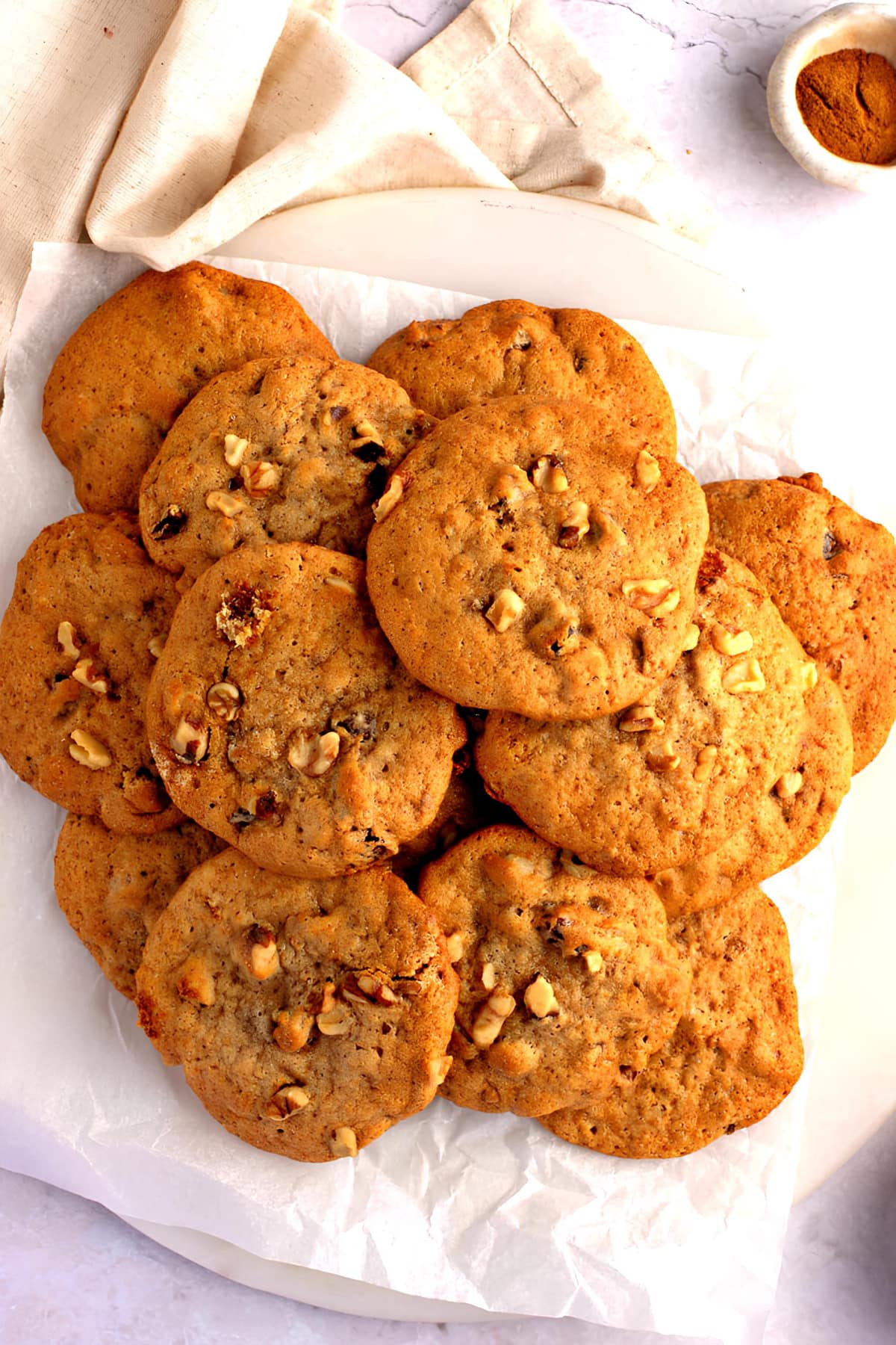 Pile of nutty and fruity persimmon cookies with walnuts and raisins on parchment paper on a plate with a bowl of cinnamon in the background