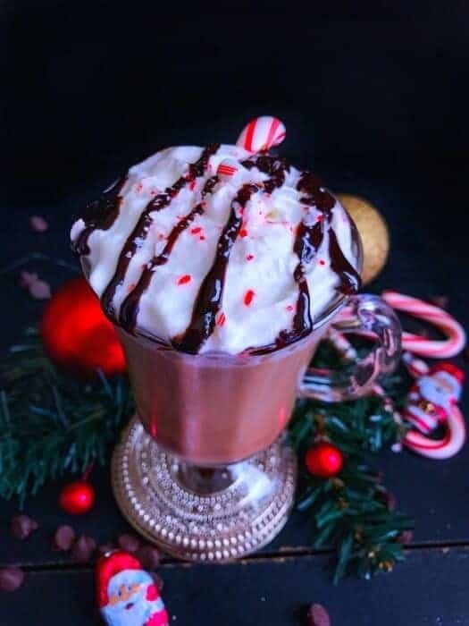 Hot chocolate on a tall glass topped with whipped cream, sprinkles and drizzle of chocolate syrup.