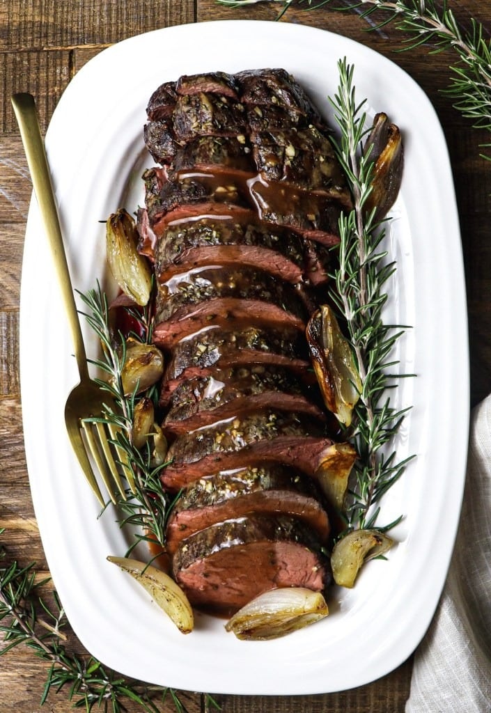 Roasted beef tenderloin sliced served with sweet roasted shallots and a wine sauce on a plate garnished with rosemary. 