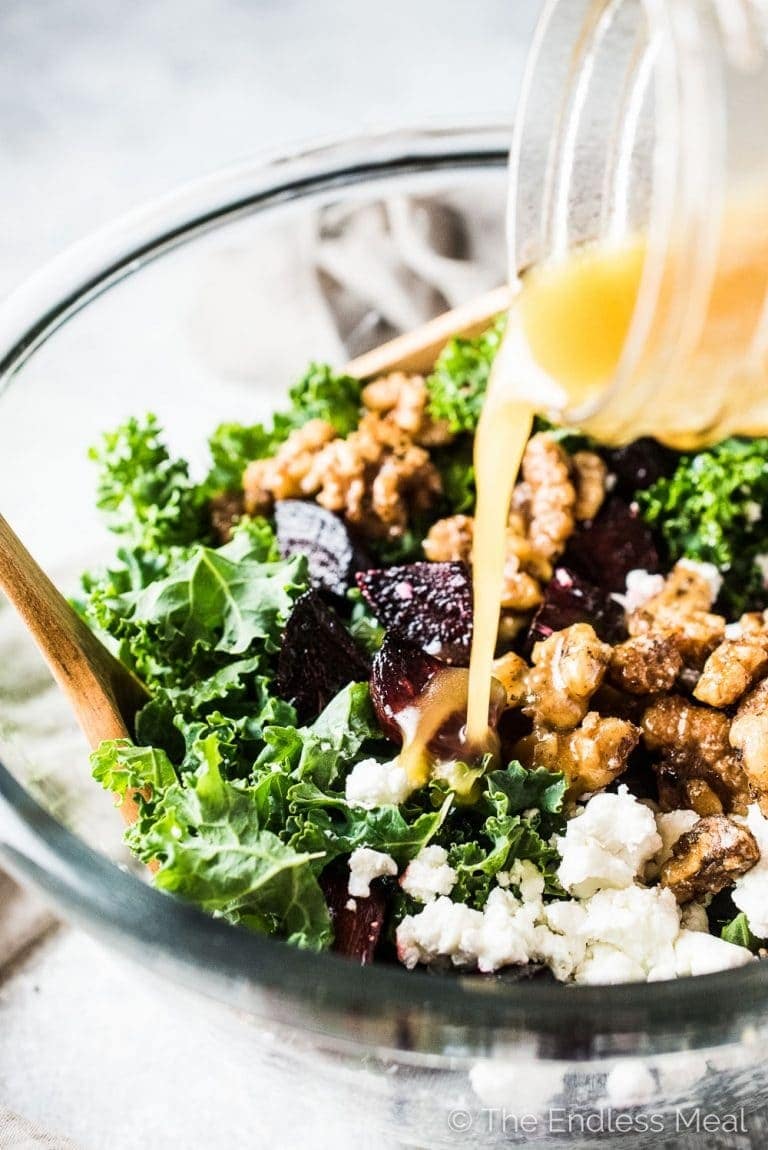 A glass of salad dressing pouring on a bowl of salad made with kale, roasted beets, maple candied walnuts, and goat cheese