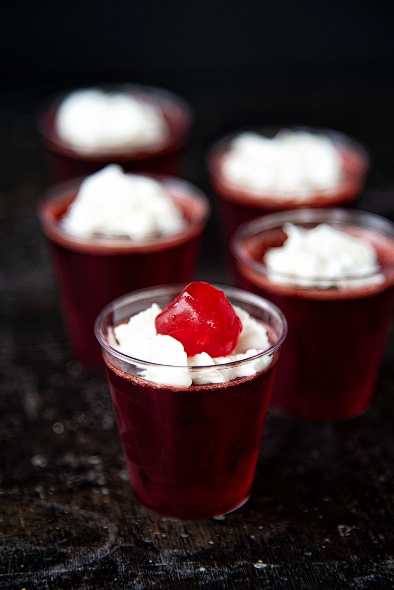 Dark red Jello shots on shot glasses topped with whipped cream and fresh cherries. 