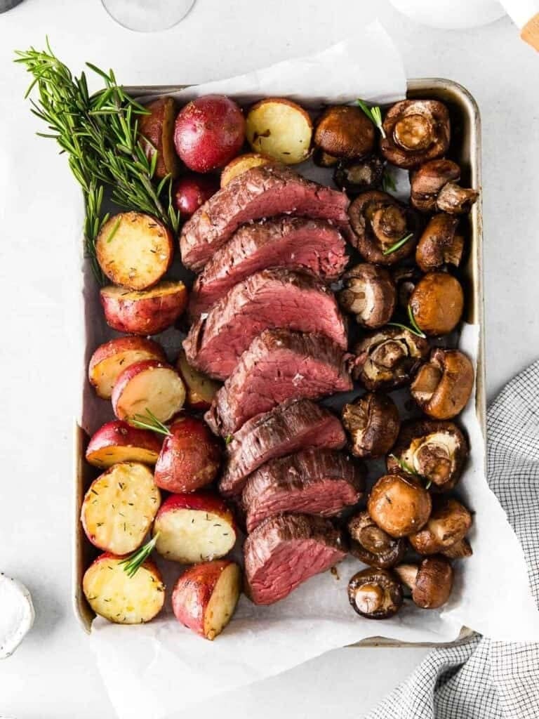 Sliced steak served on a sheet pan with roasted mushroom and potatoes.