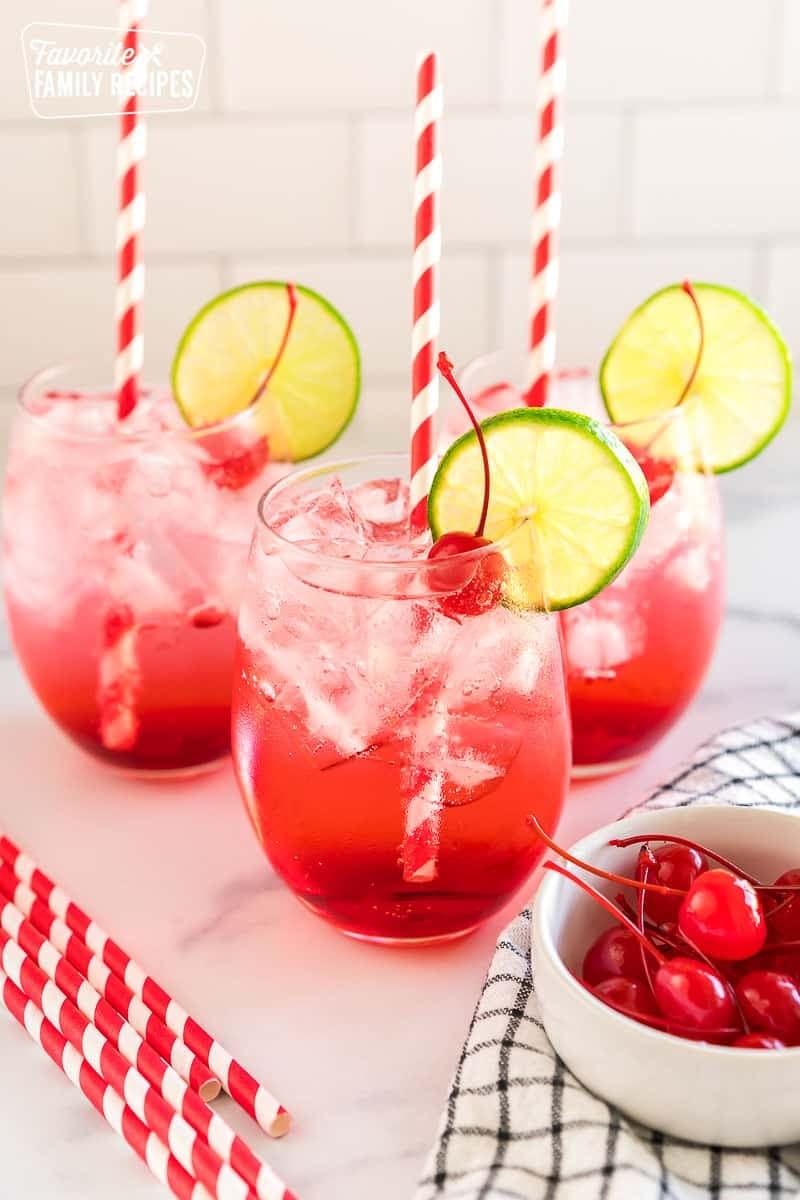 Three glasses of red colored mocktails with ice and fresh cherries garnish. 