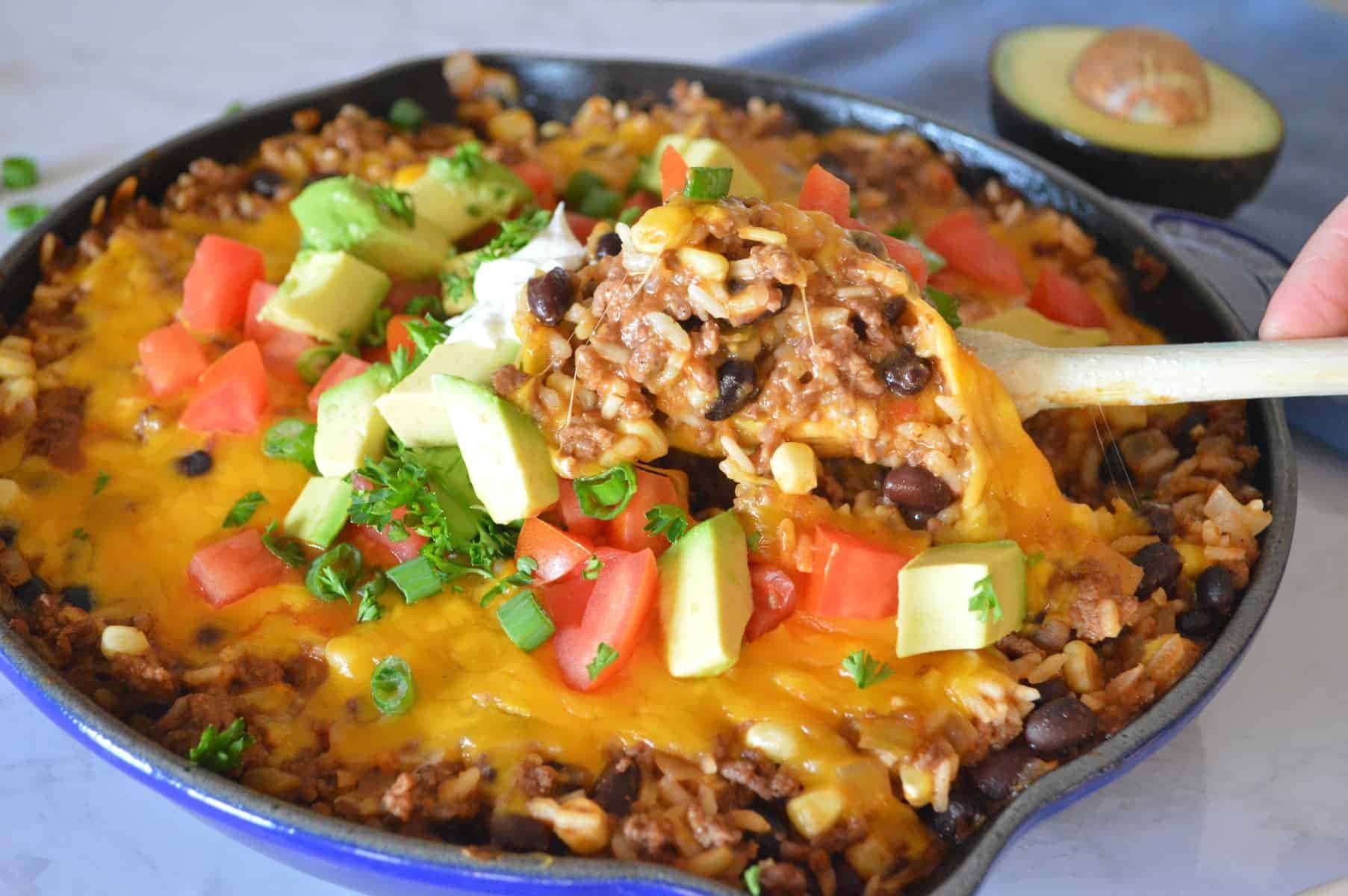 Skillet full of rice, ground beef, salsa, black beans, corn kernels and shredded cheese with chopped tomatoes, avocados and parsley