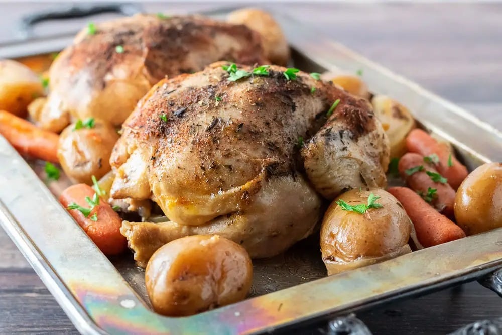 Two pieces of roasted chicken on a baking pan with potatoes, carrots and herbs.