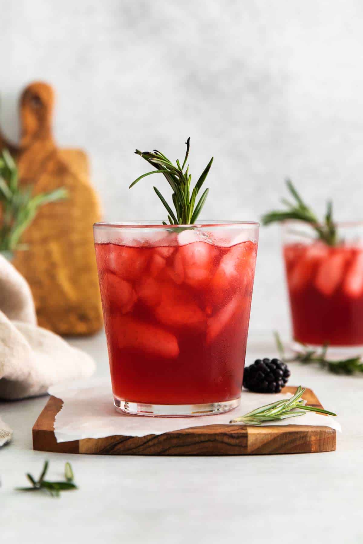 Two glasses of smoked rosemary blackberry whiskey sour cocktail garnished with rosemary, served on a wooden coaster