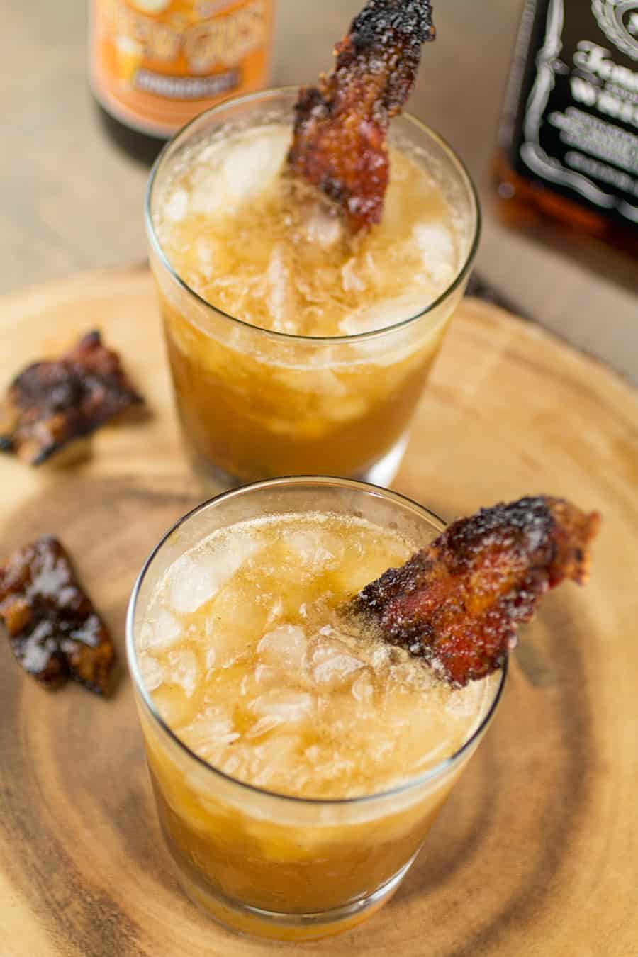 Two glasses of homemade smoky whiskey mules with candied bacon garnish on a wooden serving tray and more candied bacon around the base