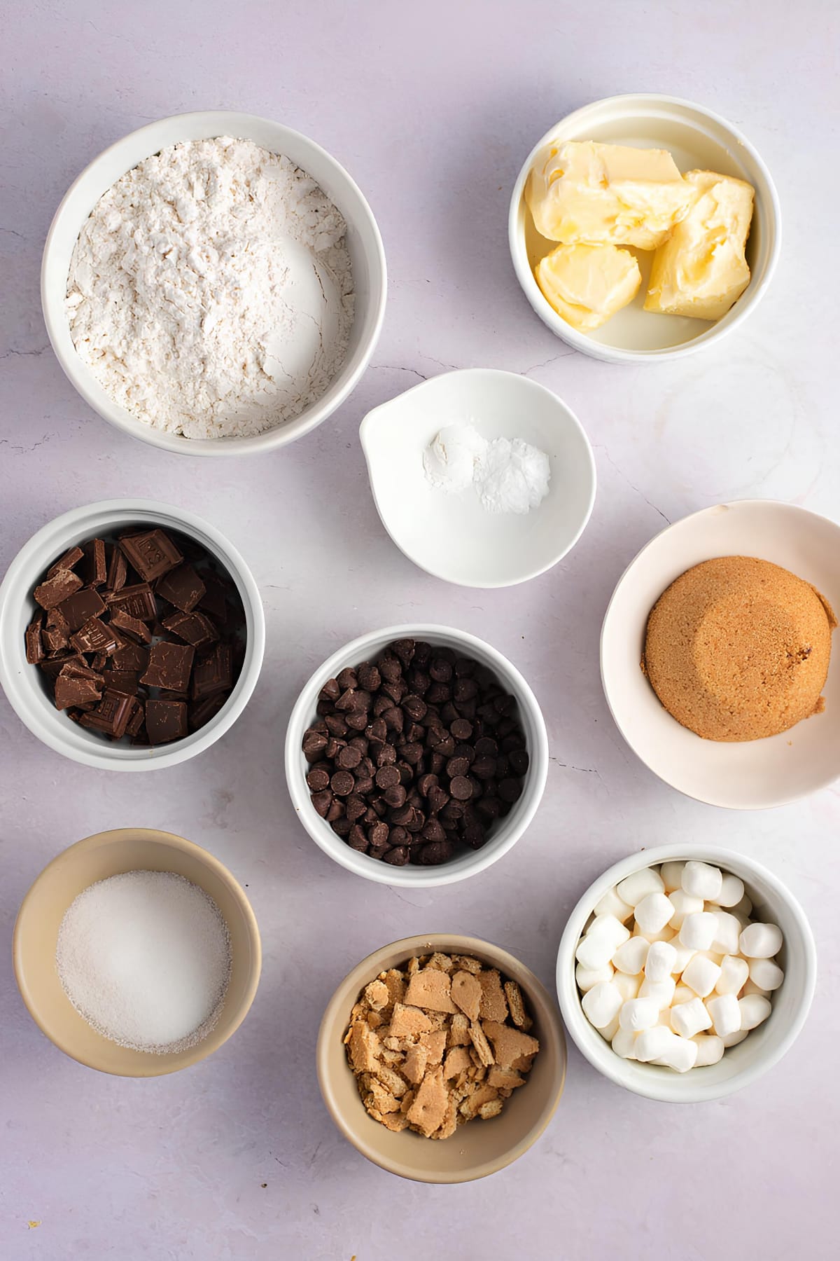 Butter, brown sugar
,flour, chocolate chips, marshmallows, Hershey's bars and graham crackers on bowls flat lay on a white table surface.