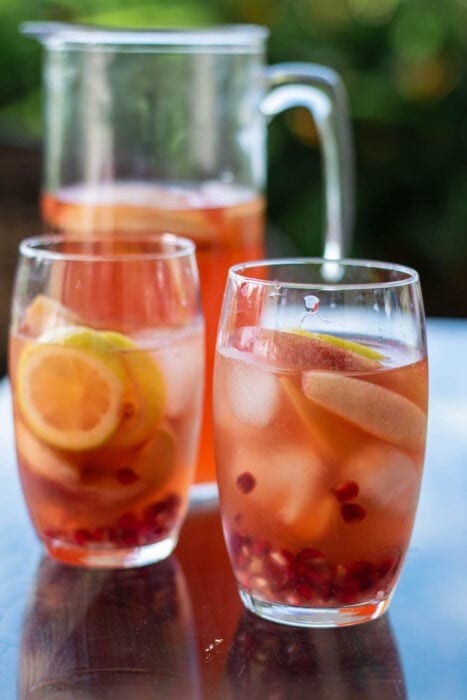 Two glasses of fruit and lemonade on a table garnished with pomegranate arils, lemon slices and peach. 