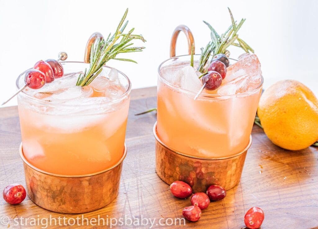 Two glasses of orange drink filled with ice garnished with rosemary sprigs and cranberries on sticks.