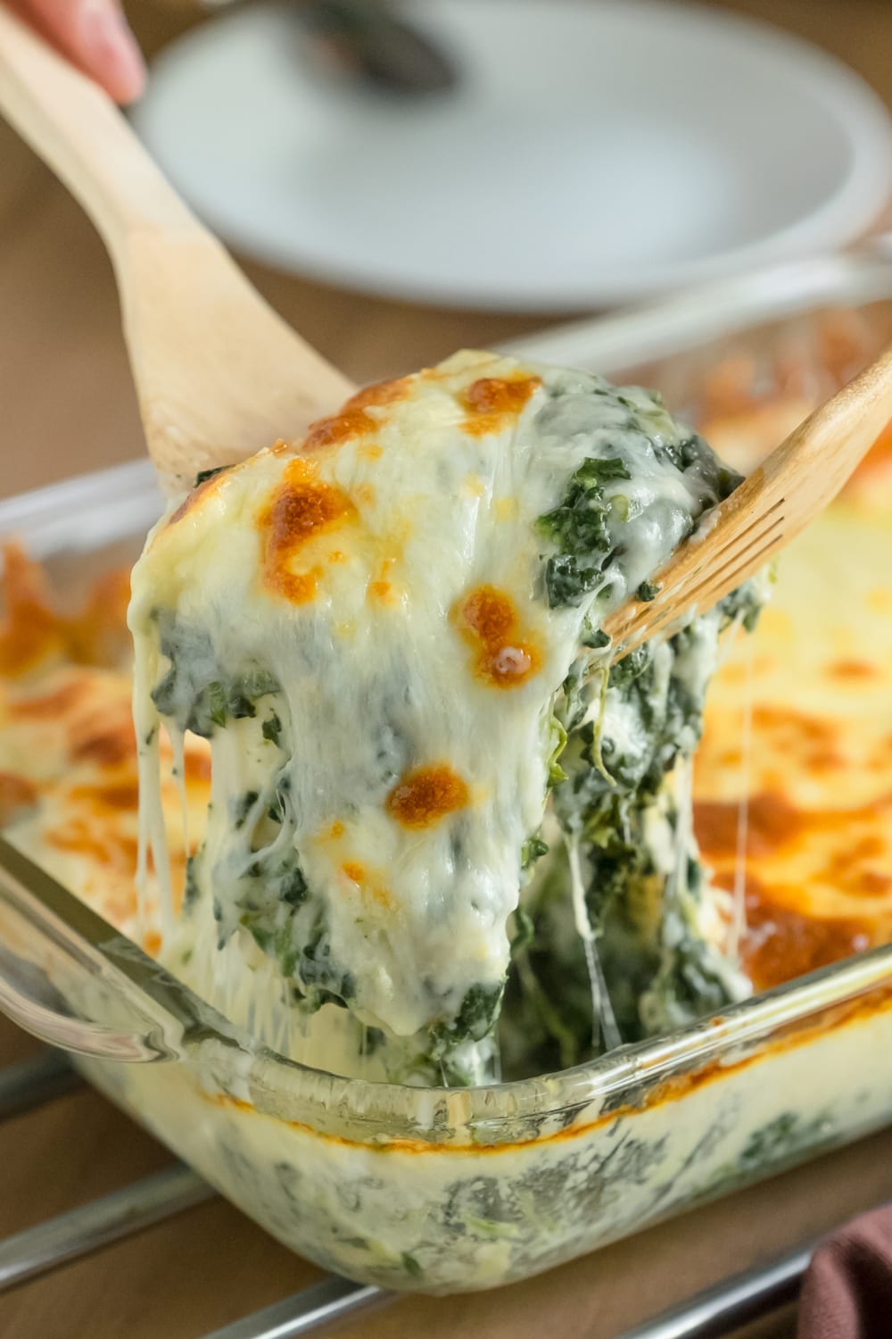 Wooden fork and spoon lifting gooey spinach artichoke dip from a glass dish.