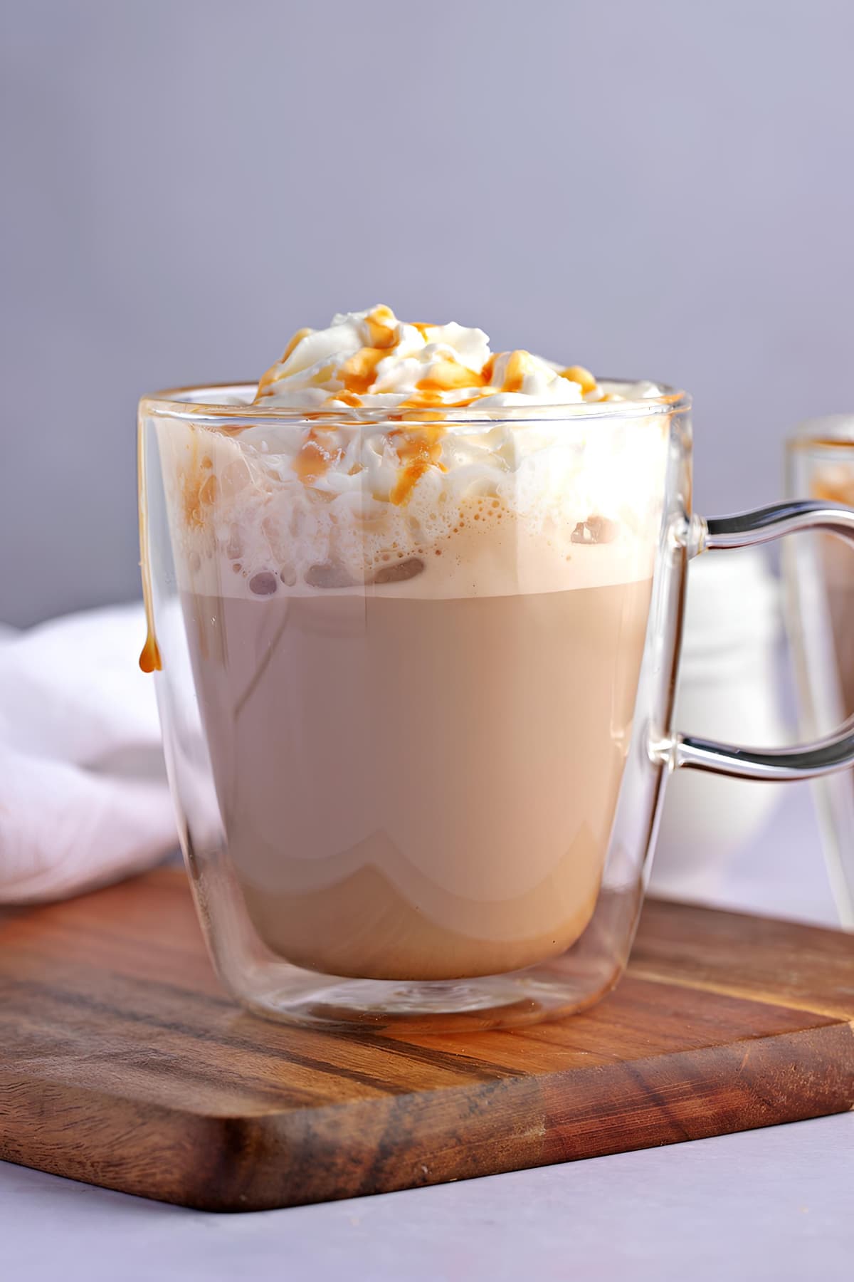 A glass mug with homemade caramel macchiato topped with whipped cream and drizzle of caramel syrup on a wooden cutting board- close up side view.