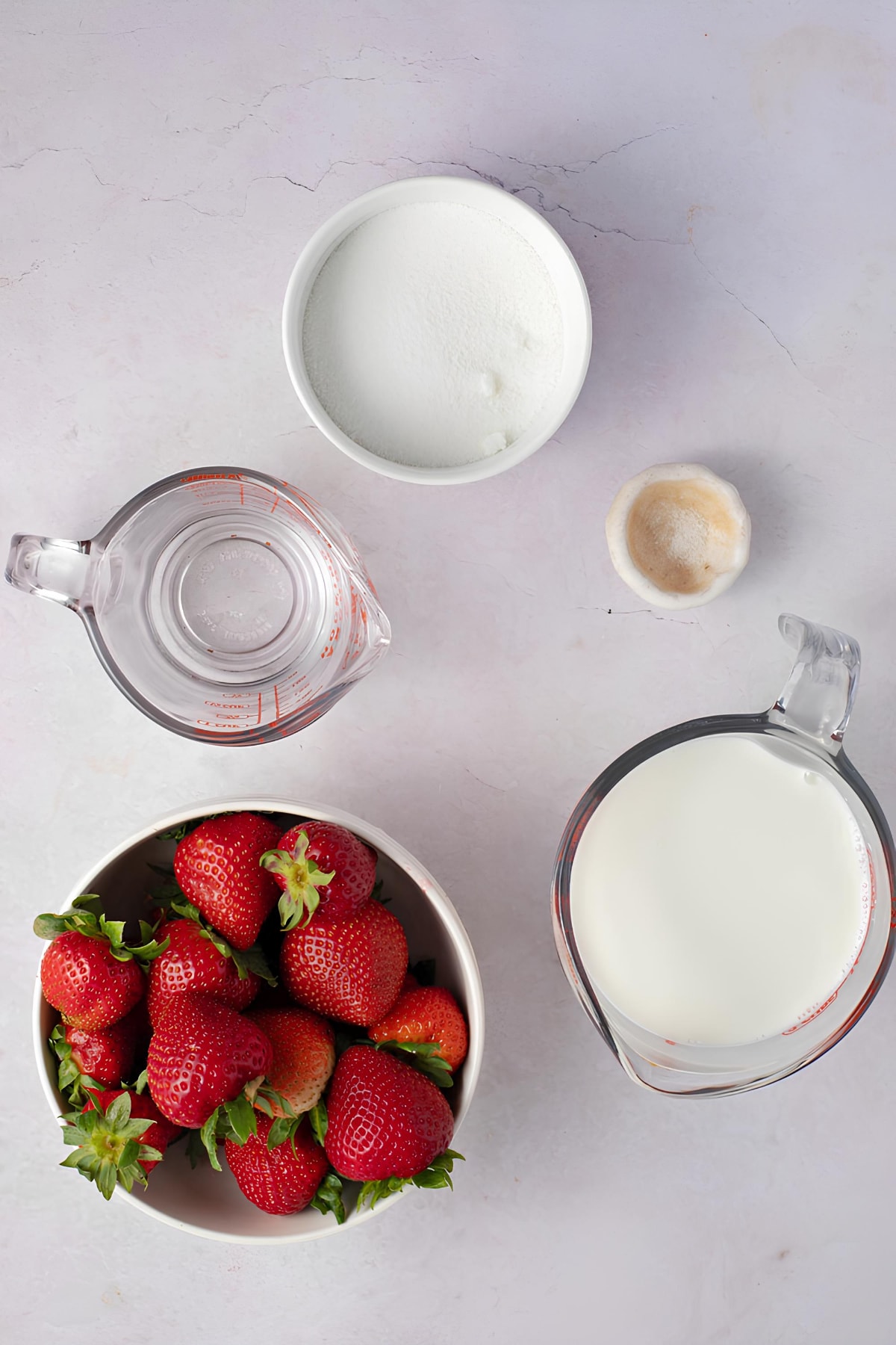 Strawberry milk ingredients on a white table including fresh strawberries, milk, sugar and whipped cream.