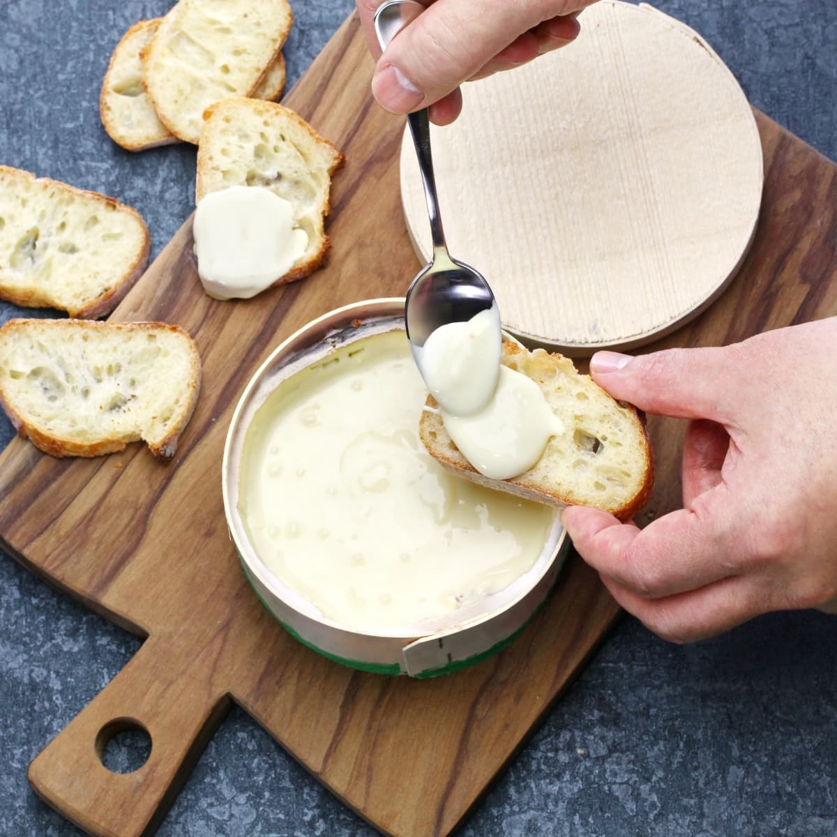 A Person Holding a Spoon over a Bowl of Melted Vacherin Cheese, Spooning Cheese onto Bread over a Cutting Board