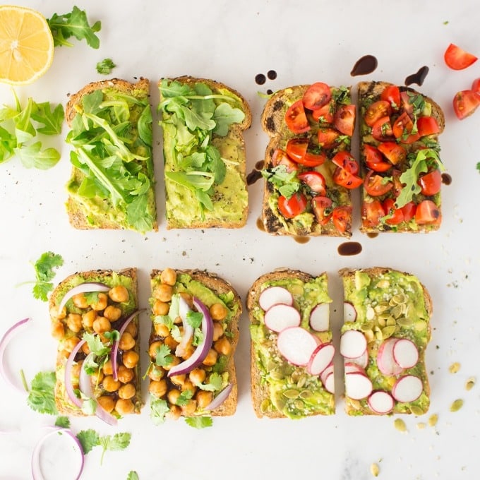 Sliced avocado toast on a table topped with different veggies like chickpeas, tomato , arugula and radish.
