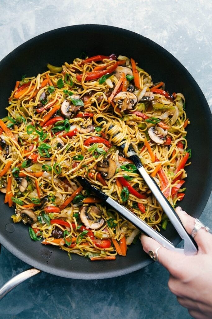 Stir fried noodles with cabbage, carrots, celery, red pepper, mushrooms, green onions and sauce tossed by a tong on a pan.