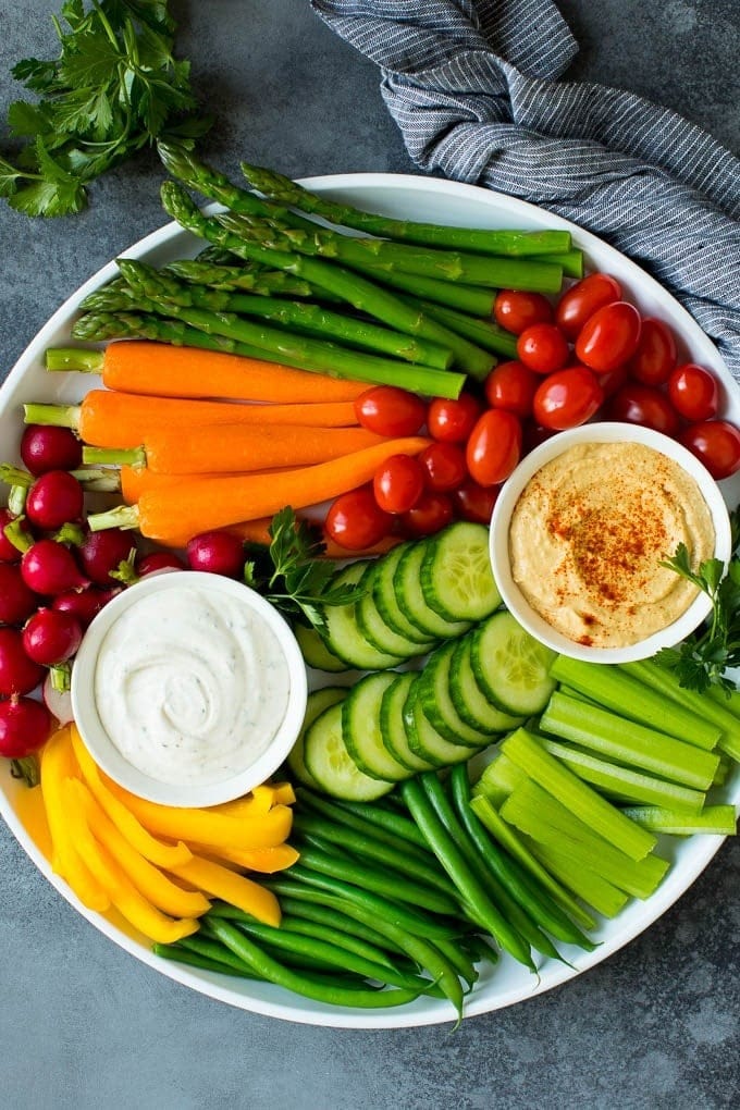 A plate full of sliced cucumber, celery, baby carrots, asparagus, berries, cherry tomatoes, hummus and mayo dip. 