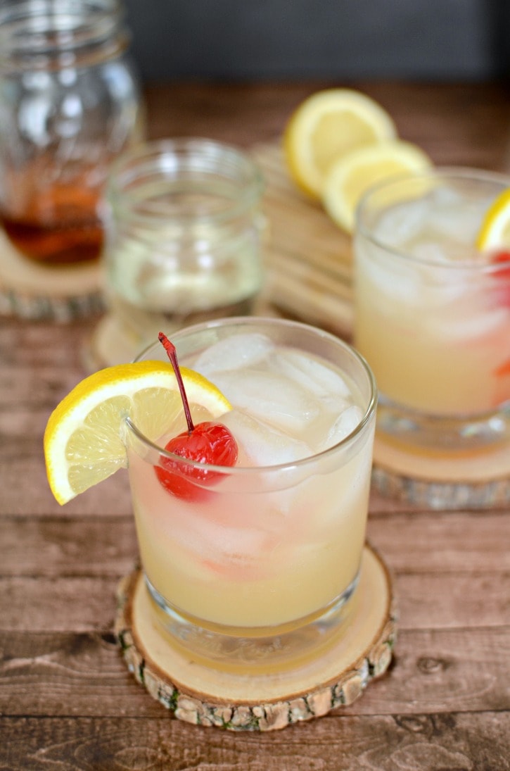 Two homemade classic whiskey sours with cherry and lemon on wooden tree coasters