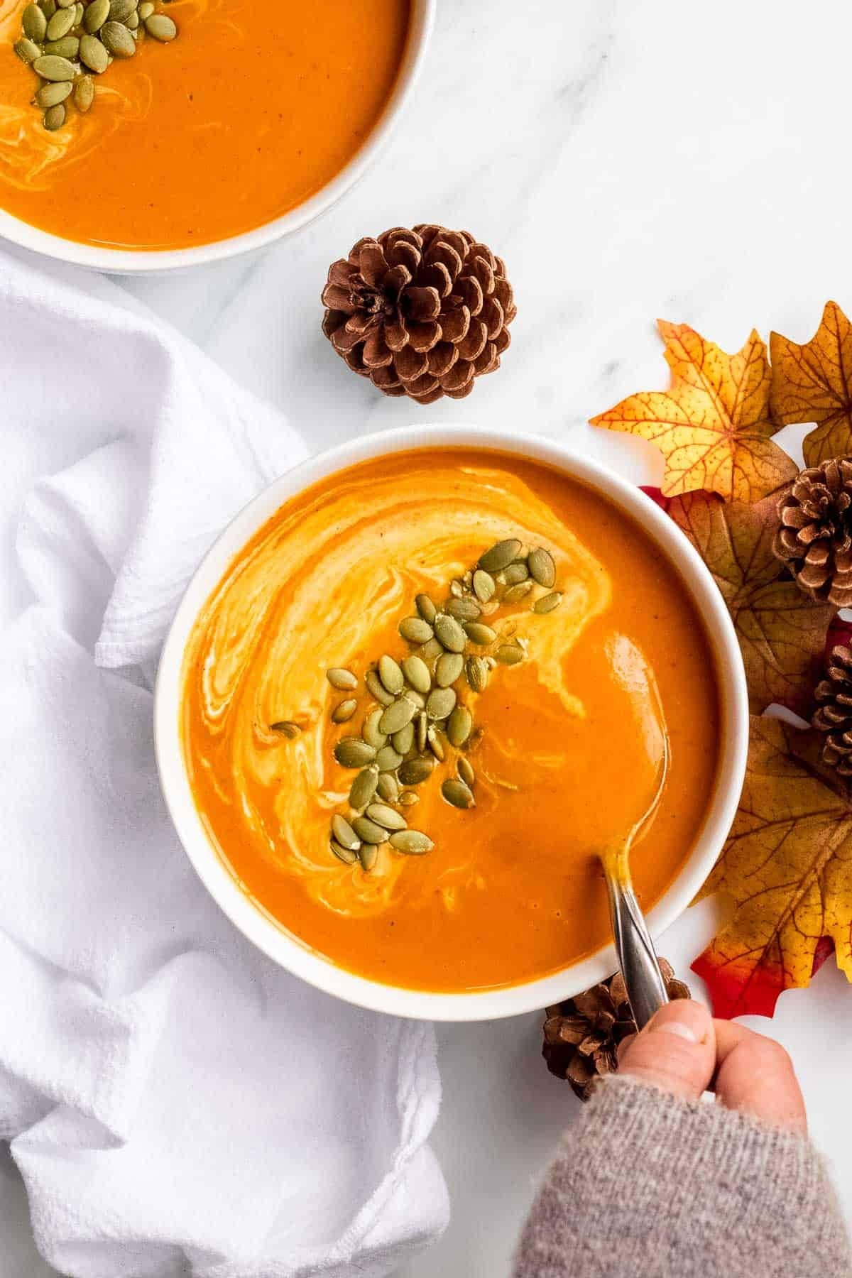 Creamy pumpkin curry soup with pumpkin seeds in a bowl with a hand about to take a spoonful- top view