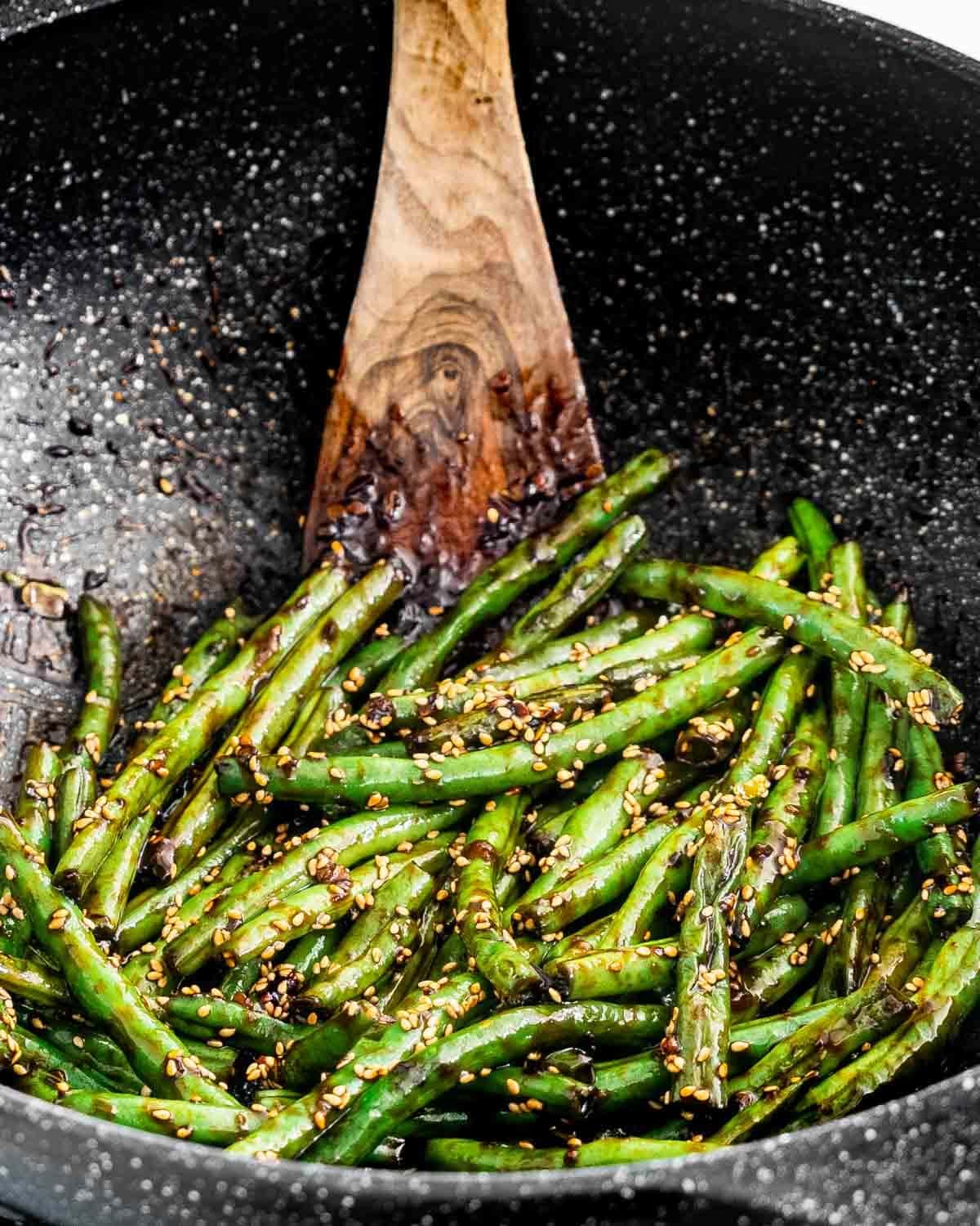 Green beans sauteed on a stone pan tossed with a wooden ladle. 