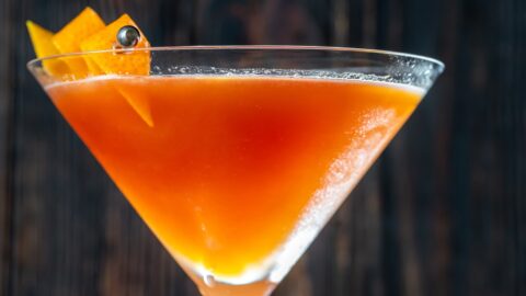 Closeup of the top of a martini glass with blood and sand cocktail garnished with orange peel on skewers.