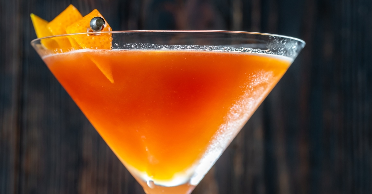 Closeup of the top of a martini glass with blood and sand cocktail garnished with orange peel on skewers.