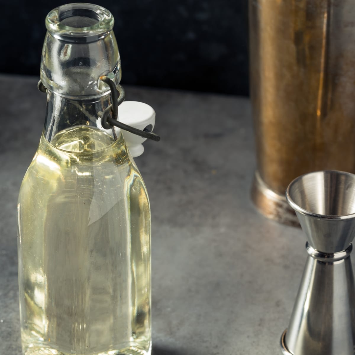 A glass bottle filled with simple syrup beside a jigger and shaker on a concrete table.
