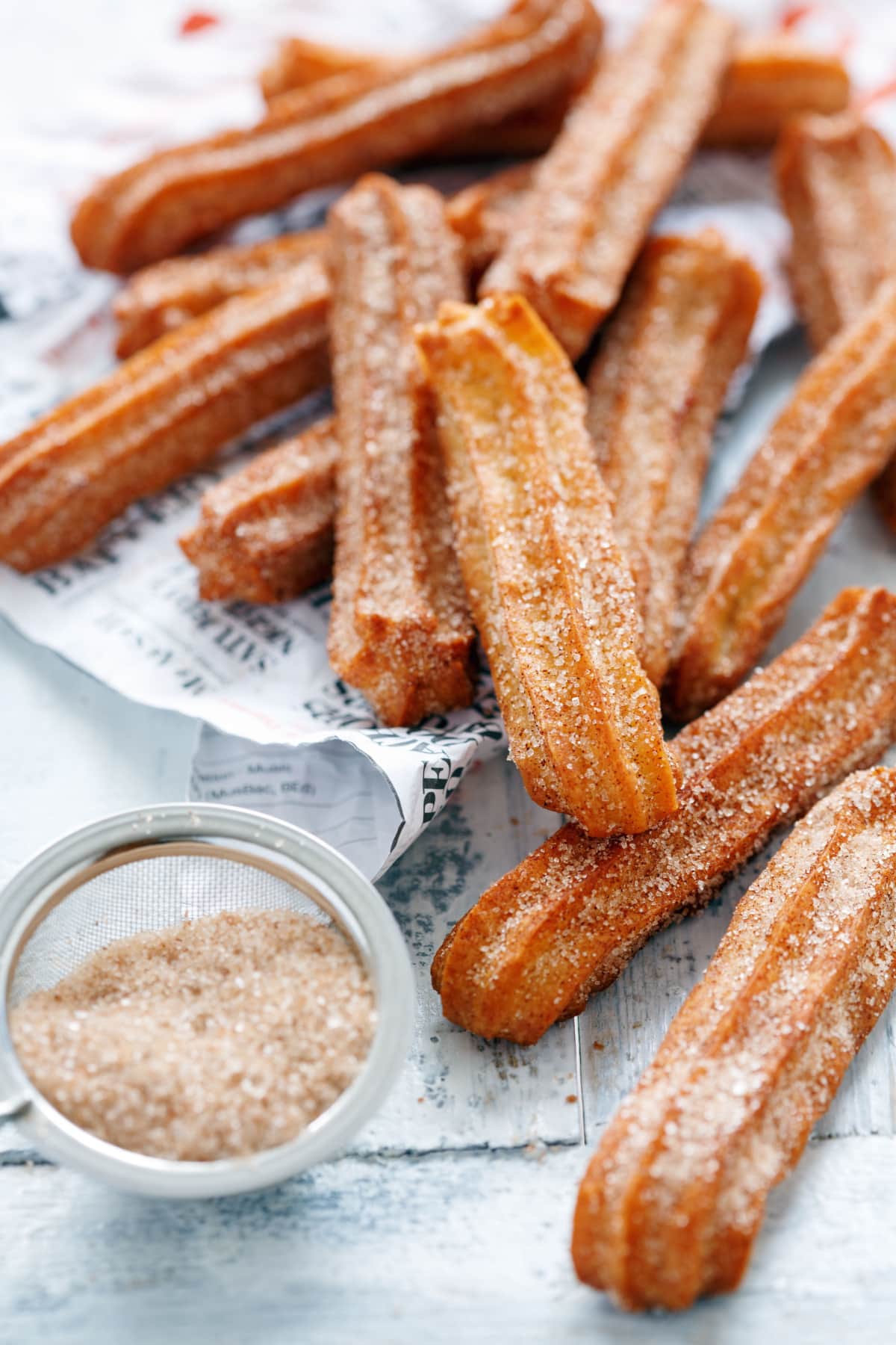 Easy Homemade Churros (Best Recipe!) featuring pile of churros sprinkled with cinnamon sugar mixture and more cinnamon sugar in a small sieve