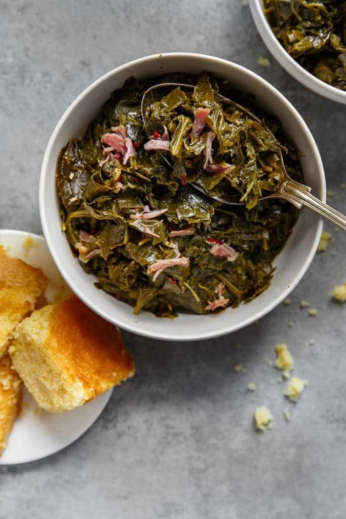 A bowl of braised collard greens on a bowl with a plate of bread rolls on side.