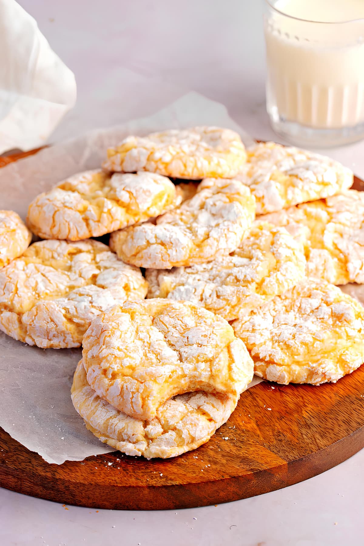 Bunch of crumbly yellow cookies on a round wooden board with a glass of milk on the background. 