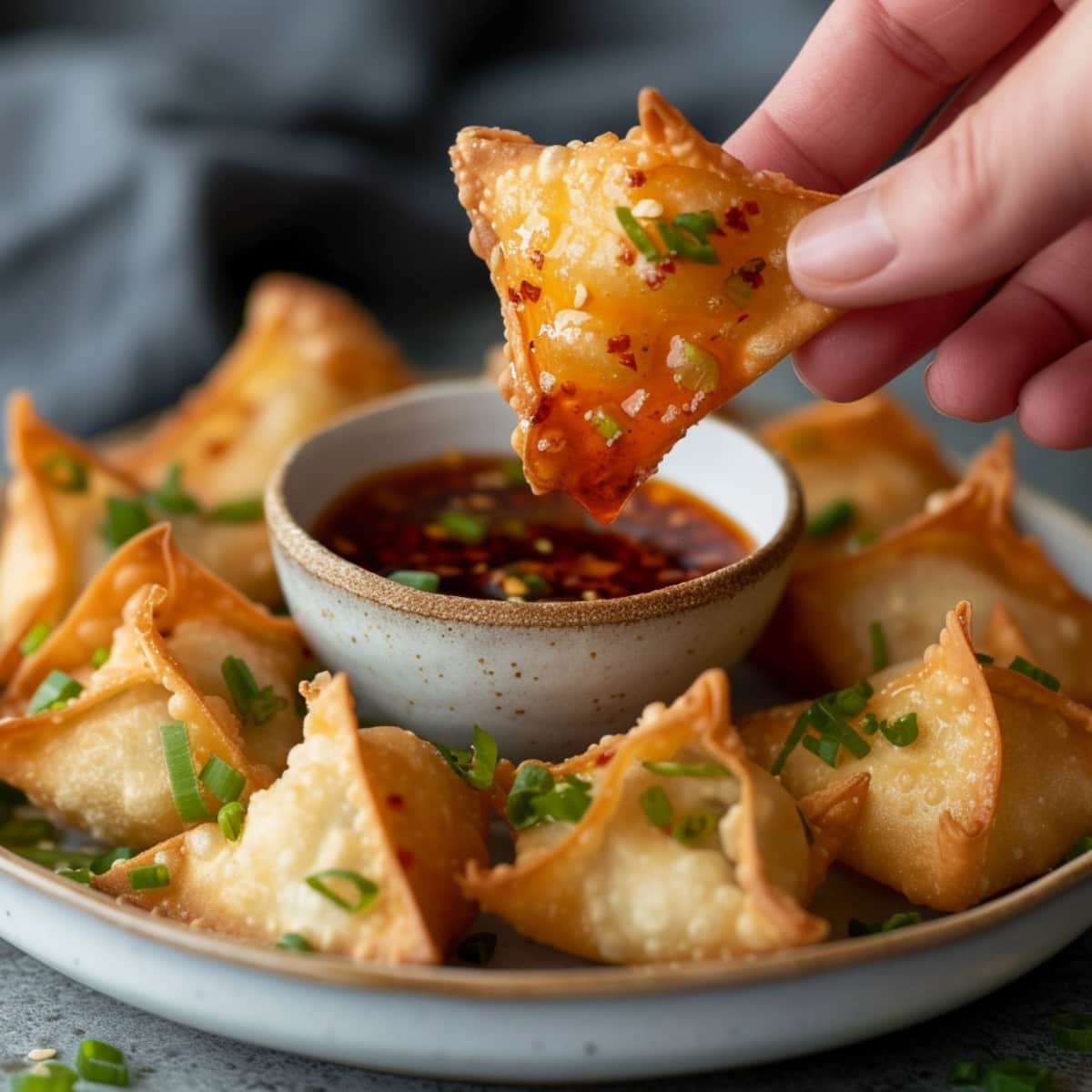 A hand dipping a crab rangoon into a bowl of sweet chili sauce