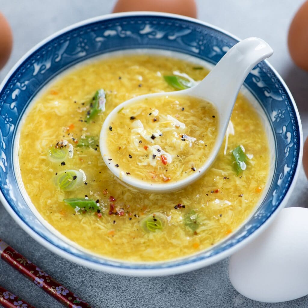 Egg drop soup served in a blue Chinese bowl with soup spoon garnished with chopped onions. 
