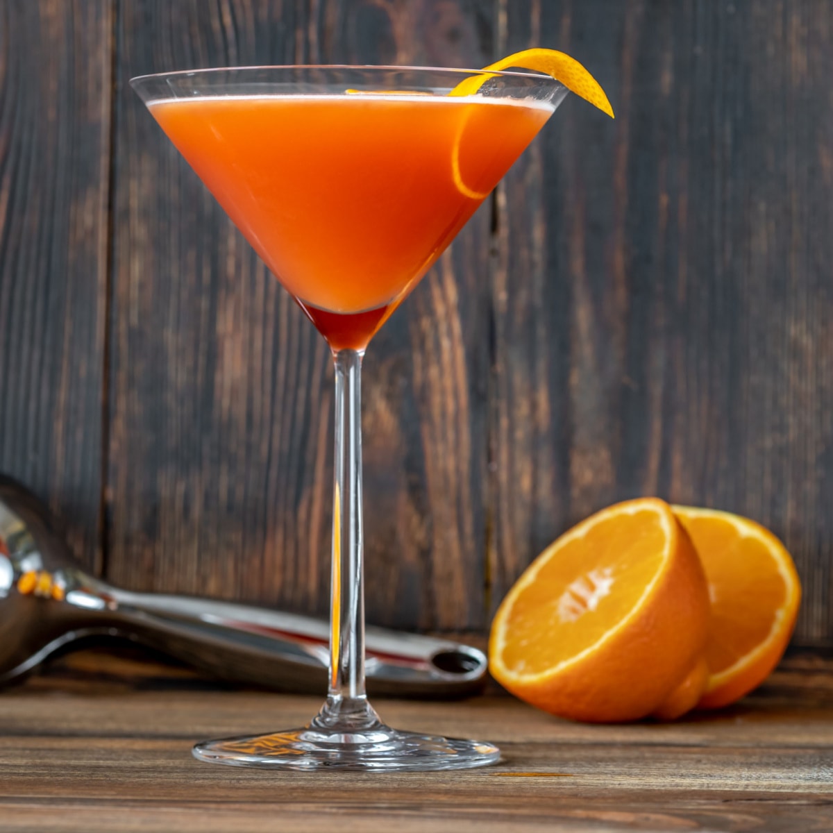 A martini glass with blood and sand cocktail garnished with orange peel beside an orange sliced in half on top of a wooden table.
