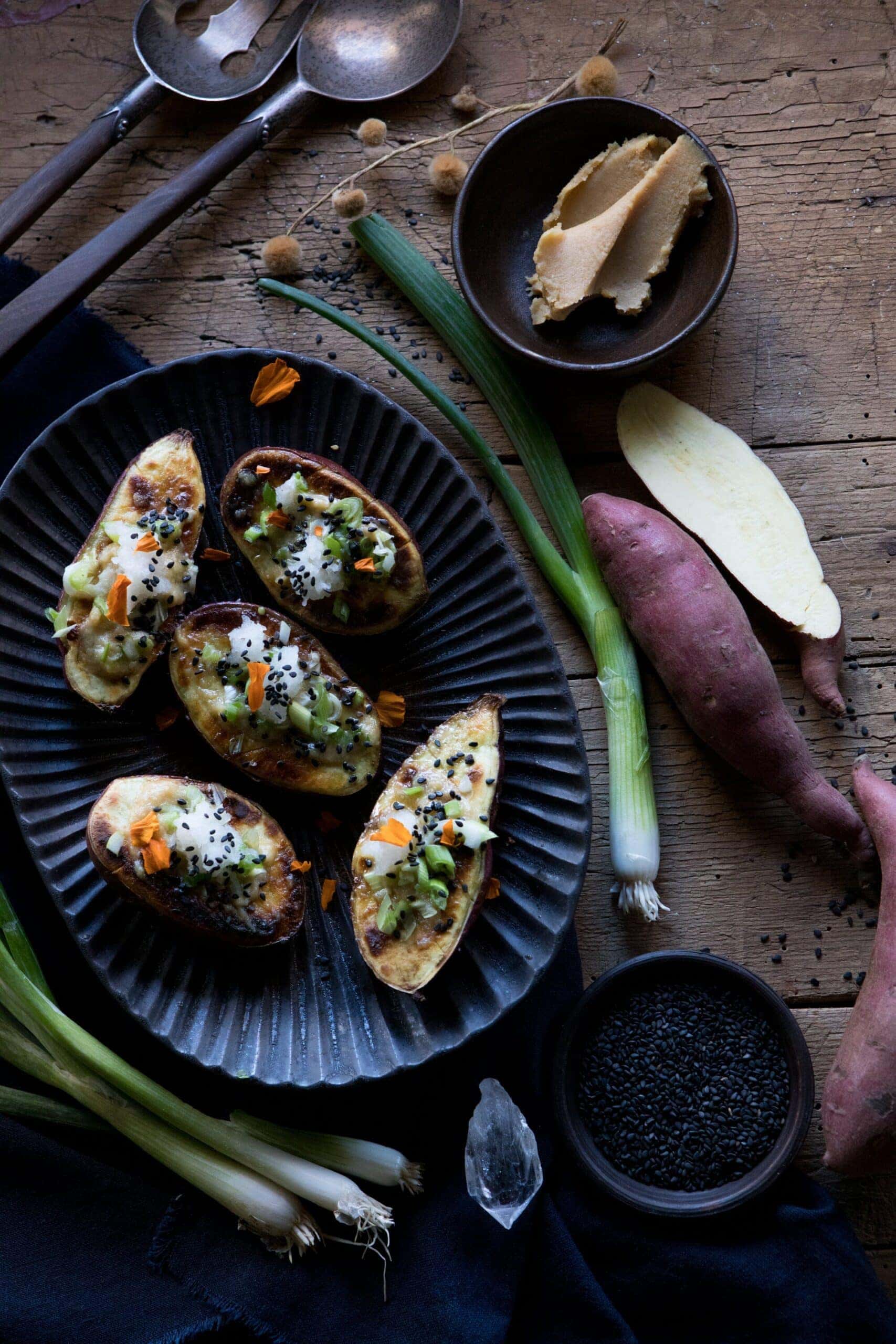 Japanese sweet potatoes with miso butter, chopped scallions and sesame seeds on a platter with raw scallions and potatoes on the table around the platter- top view