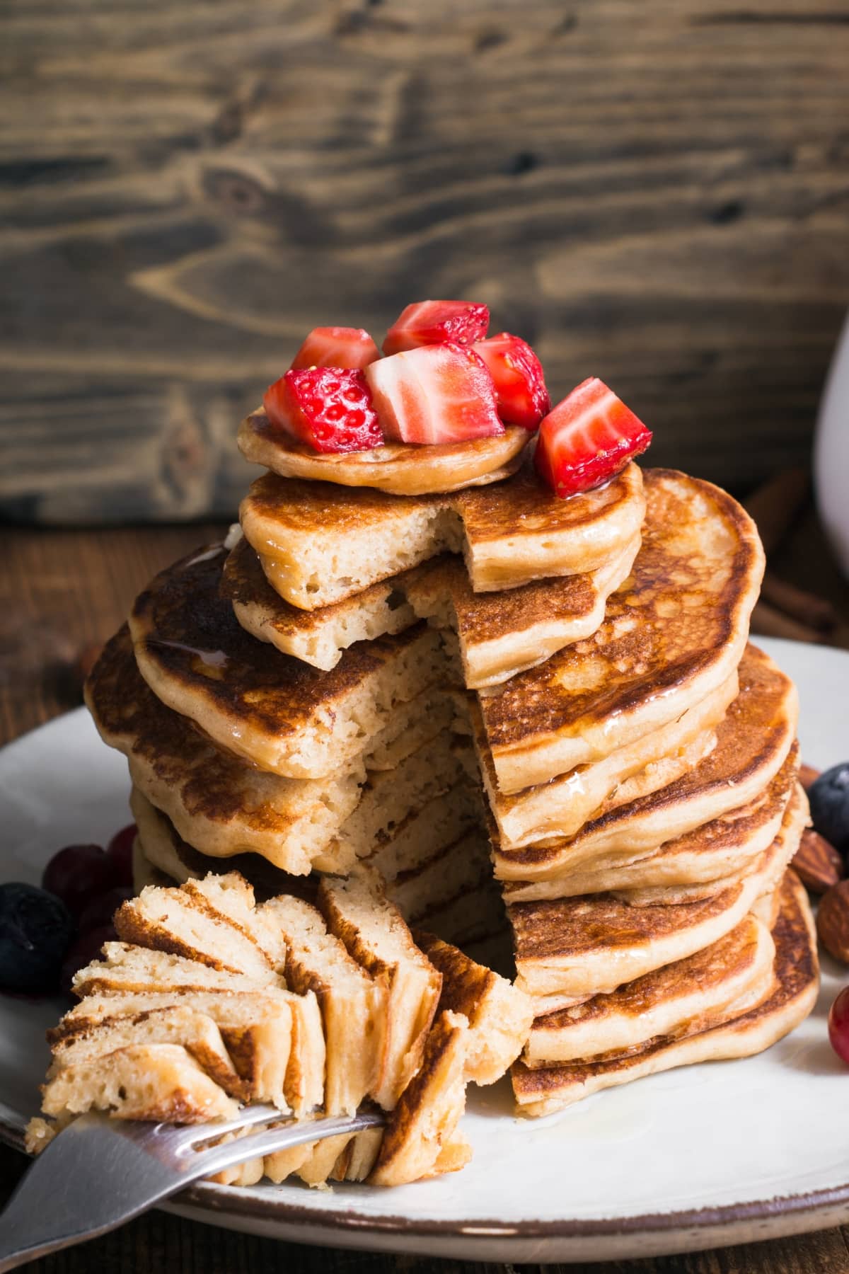 Stack of pancakes topped with slices of strawberries and maple syrup served on a white place. 