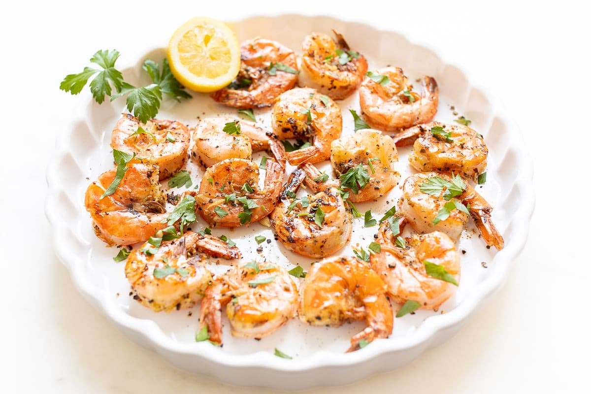 Buttered shrimp seasoned with pepper and lemon garnished with chopped parsley served on a round plate.