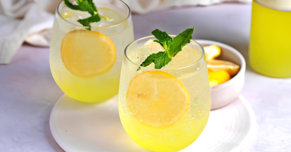 Glass filled with iced limoncello spritz cocktail garnished with lemon slices and mint leaves served on a round tray.
