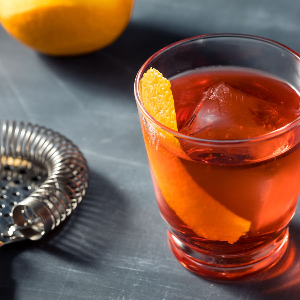 A serving of Mezcal Negroni cocktail in a glass with ice garnished with lemon peel and strainer on the side. 