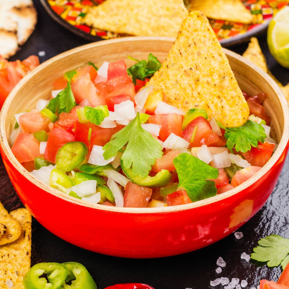A red bowl with chopped tomatoes, onions, cilantro, and jalapenos served with nacho chips.
