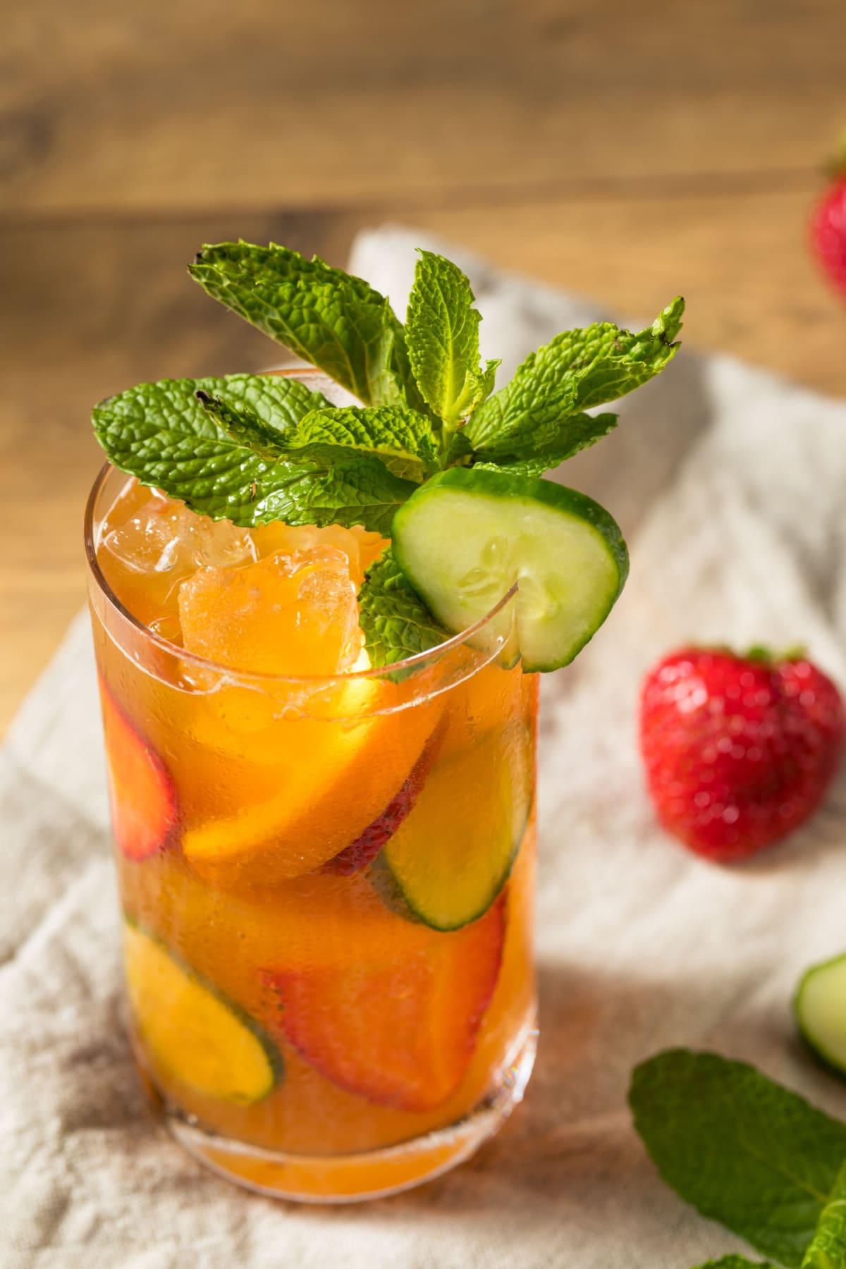Pimm’s Cup Cocktail (Classic British Recipe) featuring Pimm's Cup cocktail in highball glass filled with ice, slices of lemon, cucumber, strawberries, and mint sprig. 