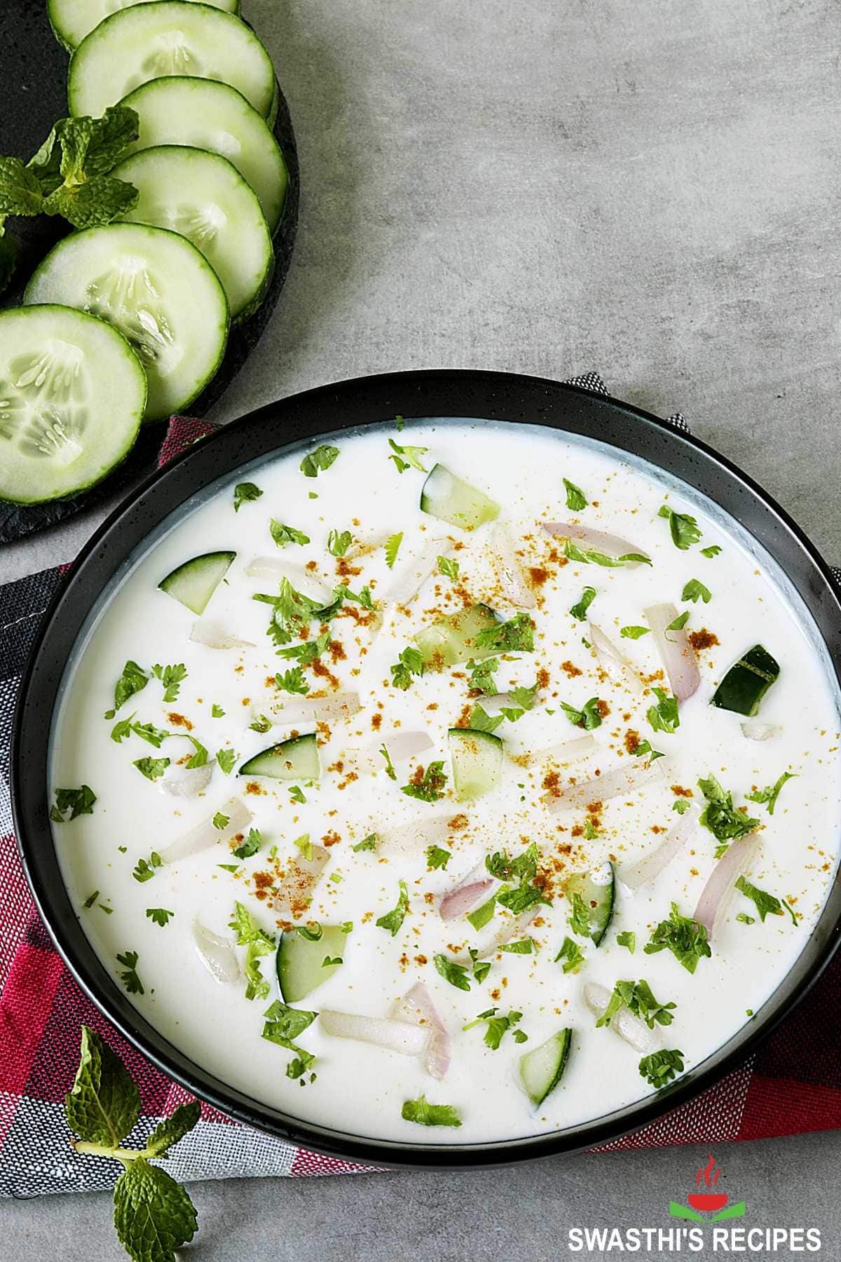 A serving of raita on a black bowl with sliced cucumber and chopped herbs.