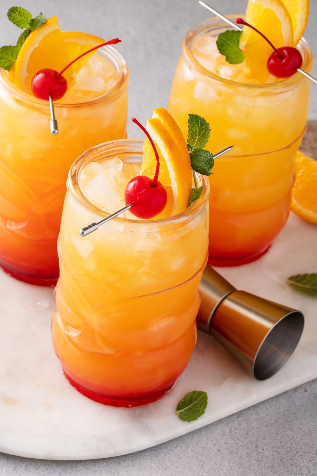 Three glasses of rum punch on tiki glasses filled with ice garnished with cherries and orange slice. 
